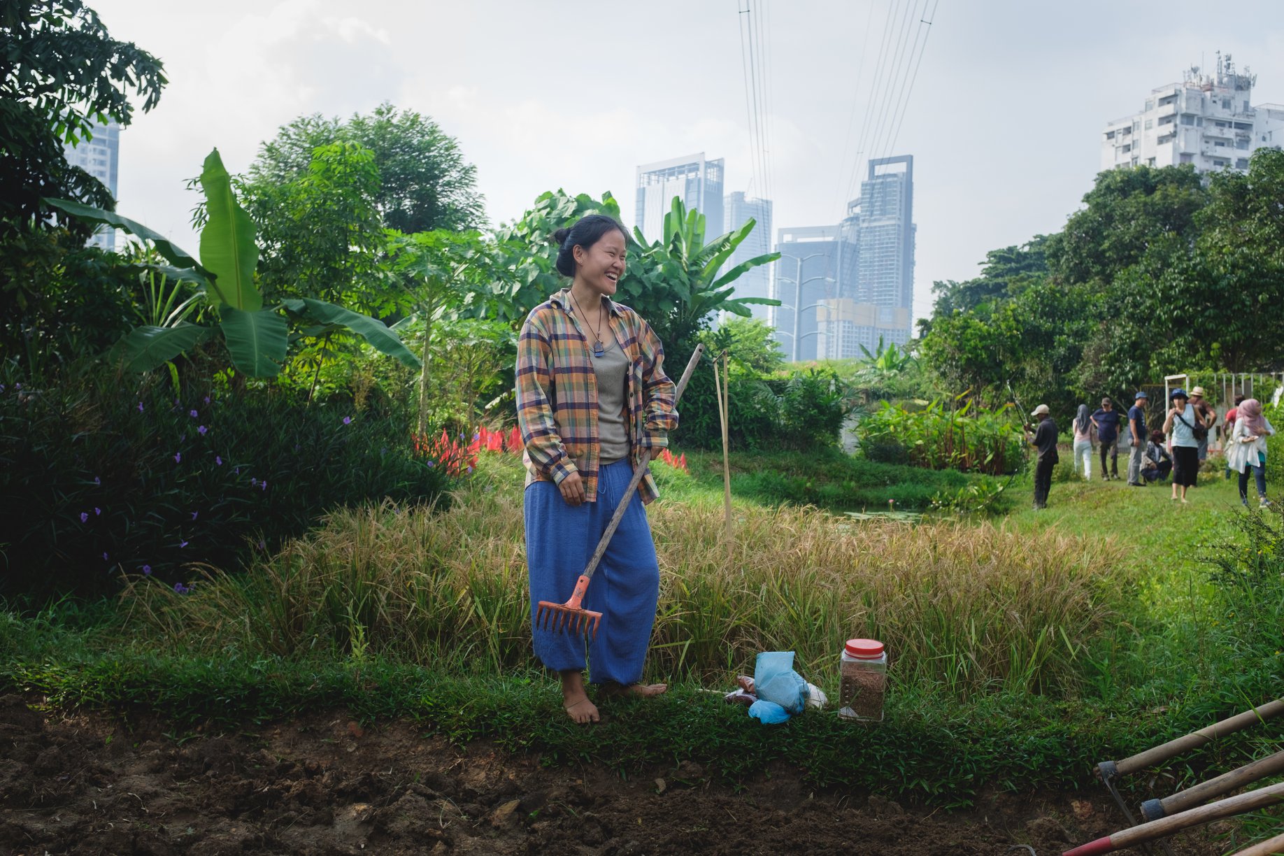 Ecologist and herbalist Khimjoe Lim is standing in a lush garden in the centre of the city in Kuala Lumpur. She is laughing at something/someone out of sight to the right of the frame and holding a rake.